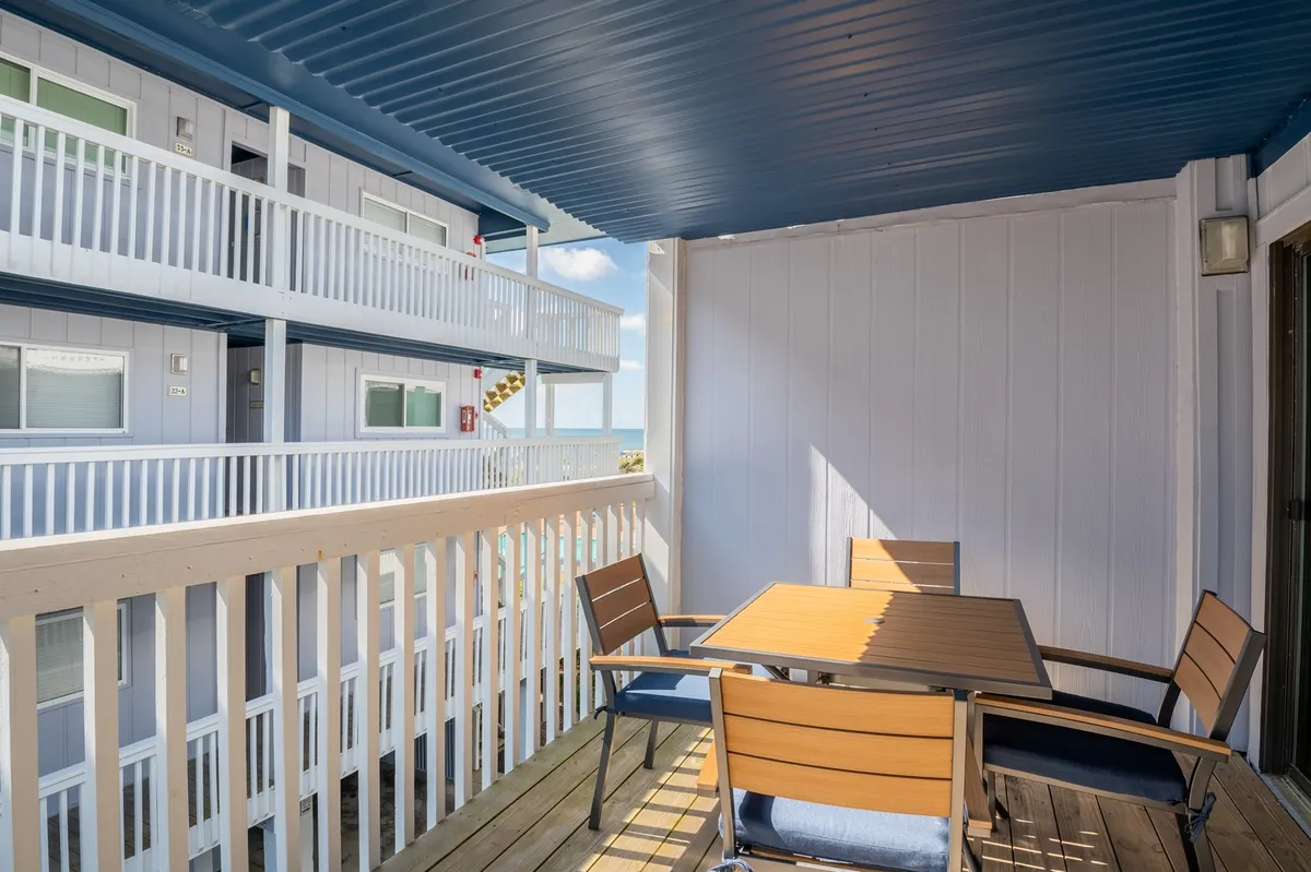A cozy yet spacious balcony area with a table and chairs arranged for casual dining or social gatherings. The blue ceiling adds a coastal touch, and the open railings frame glimpses of the outdoor scenery, evoking a breezy, seaside feel.