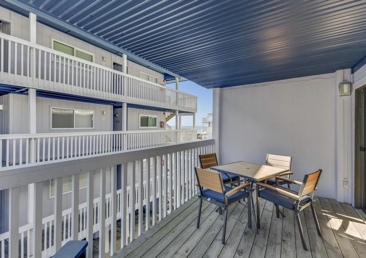 A spacious balcony featuring a teak table and comfortable chairs, bordered by a white railing and shaded by a blue ceiling. The setup is ideal for outdoor dining or relaxation, with space to move around and enjoy views beyond the railing.
