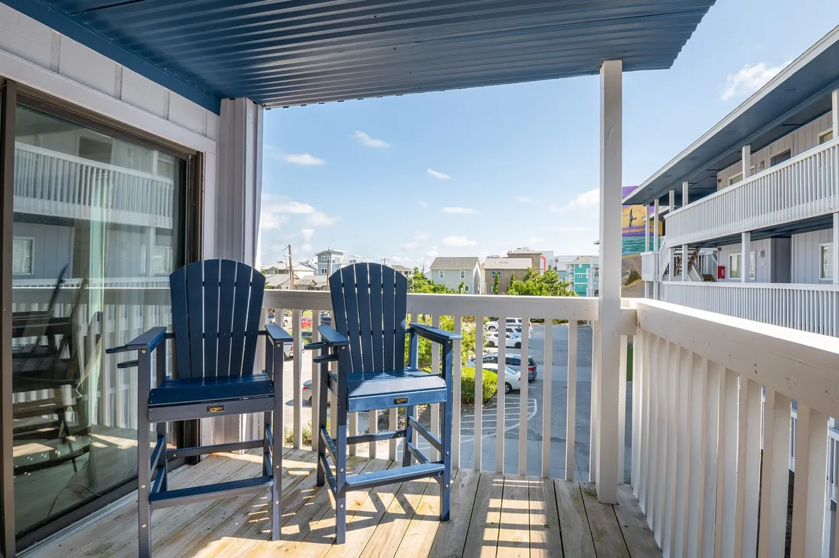This inviting exterior image shows two blue chairs arranged on a private balcony, overlooking the parking lot with partial views of the ocean beyond the railing. The scene is bathed in gentle morning light, suggesting a tranquil spot for sipping coffee while enjoying fresh ocean air.