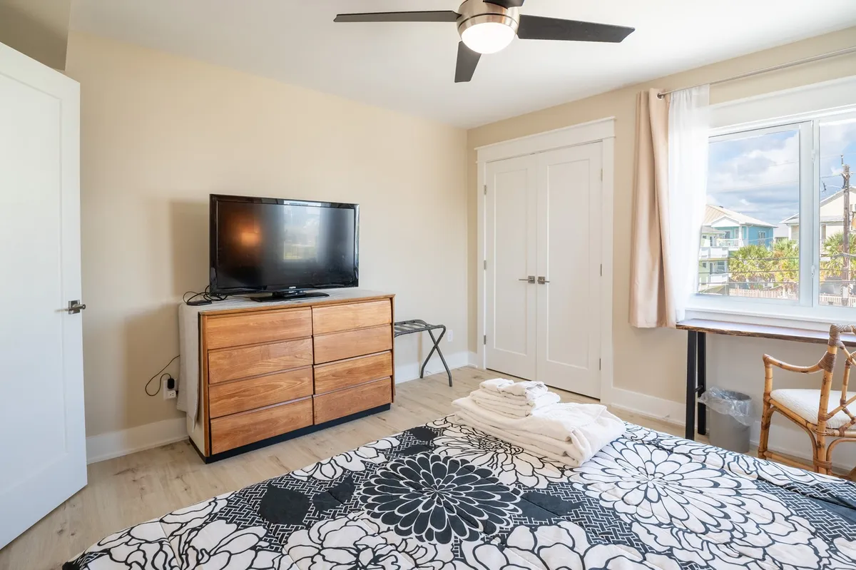 A tranquil bedroom scene featuring a bed with floral bedding, a sleek wooden dresser, a television, and a large window flooding the space with natural light. The room exudes comfort and serenity, ideal for both relaxation and productive work sessions.