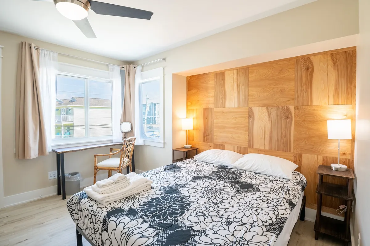 A stylish bedroom features a plush bed, warm wood panel wall, windows for natural light, a chair, and a lamp—creating a soothing environment ideal for relaxation or reading by the window.