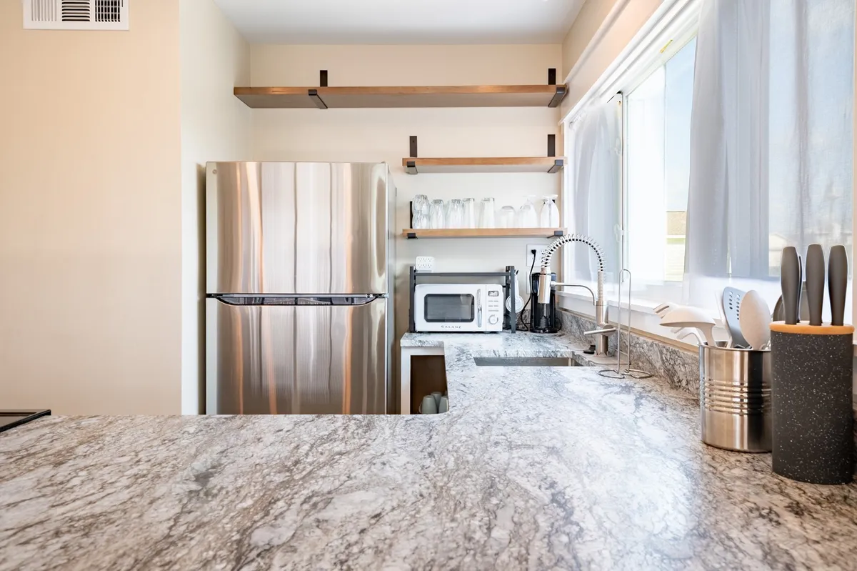 A modern kitchen featuring a sleek stainless steel refrigerator, leathered granite countertops, and open shelving with stylish glassware. The space is designed for both culinary creativity and social gathering, feeling both luxurious and functional.