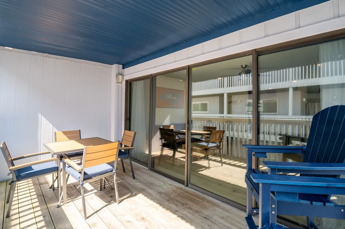 The image features a wooden deck with comfortable outdoor furniture and a blue ceiling, offering an inviting spot to relax or share meals outdoors. The casual arrangement suggests a communal area ideal for gatherings, picnics, and enjoying refreshing breezes at any time of day.