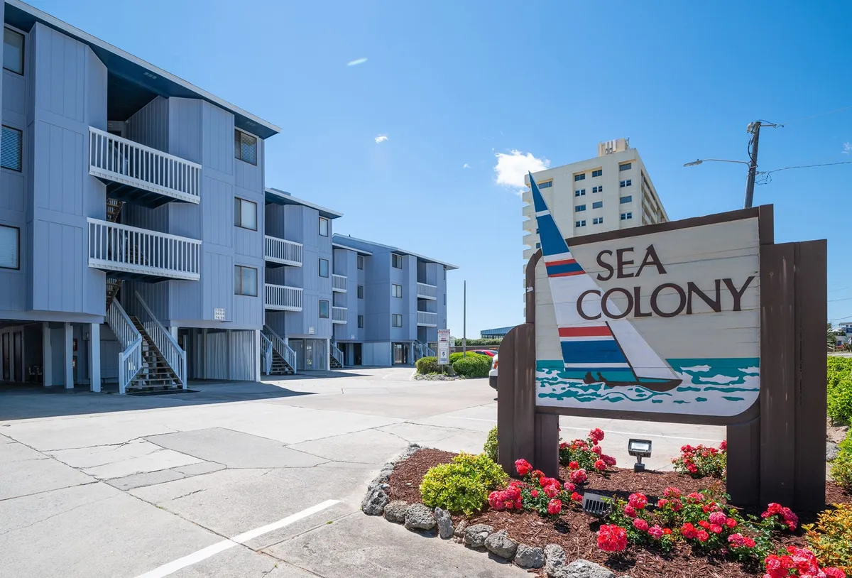 This exterior image showcases the Sea Colony entrance sign, the building’s architectural style, and vibrant flower beds, all under bright, inviting sunlight. It hints at the well-maintained grounds, ample parking, and a welcoming resort-like environment.