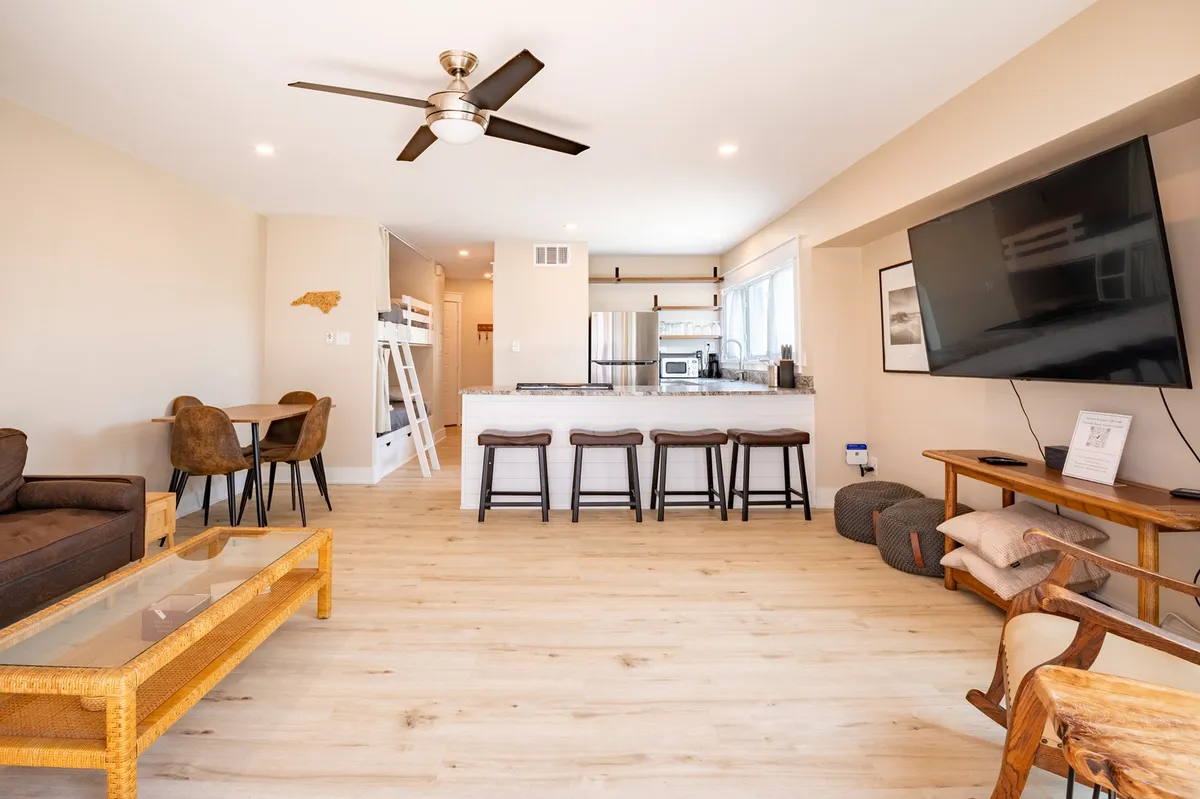 A spacious, open-concept living area seamlessly blends a comfortable sofa, large flat-screen TV, and a dining table with chairs. The modern kitchen features an island with bar stools, inviting guests to gather and enjoy casual meals. Light fills the space through expansive glass doors, enhancing the airy, contemporary feel.