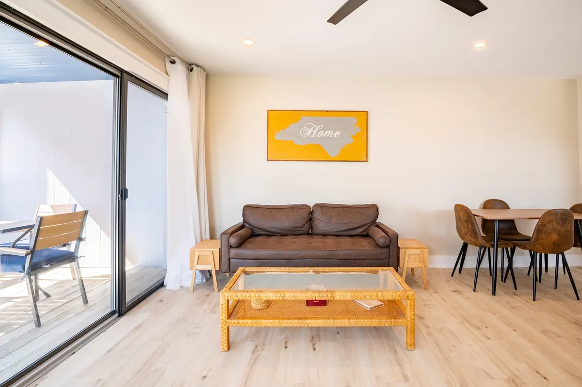 A cozy living space with a plush sofa, elegant artwork, and a dining table set near large windows. Natural light streams in, casting a warm glow over the area, and shiplap details subtly accent the walls for a coastal touch.