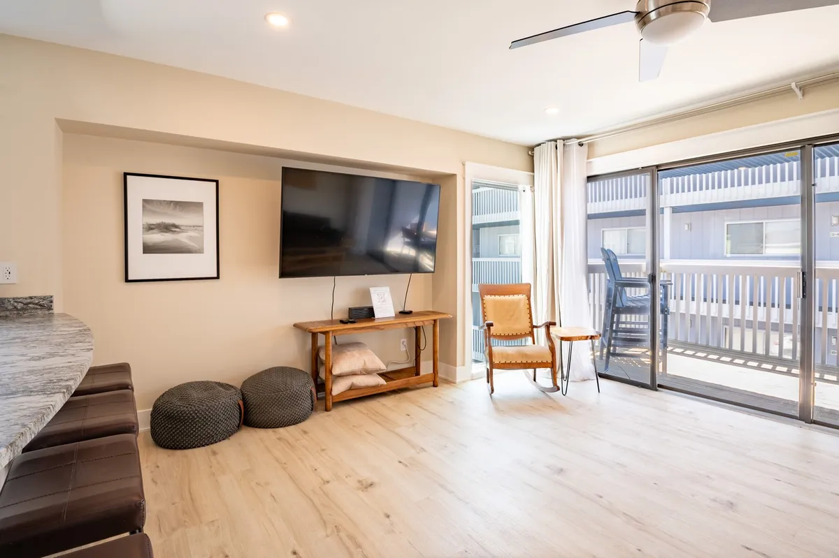 A comfortable living area with a television, wooden chair, sliding glass door, and bar stools. Sunlight streams through the door, creating an inviting space for relaxation or socializing, with easy access to the outdoors.