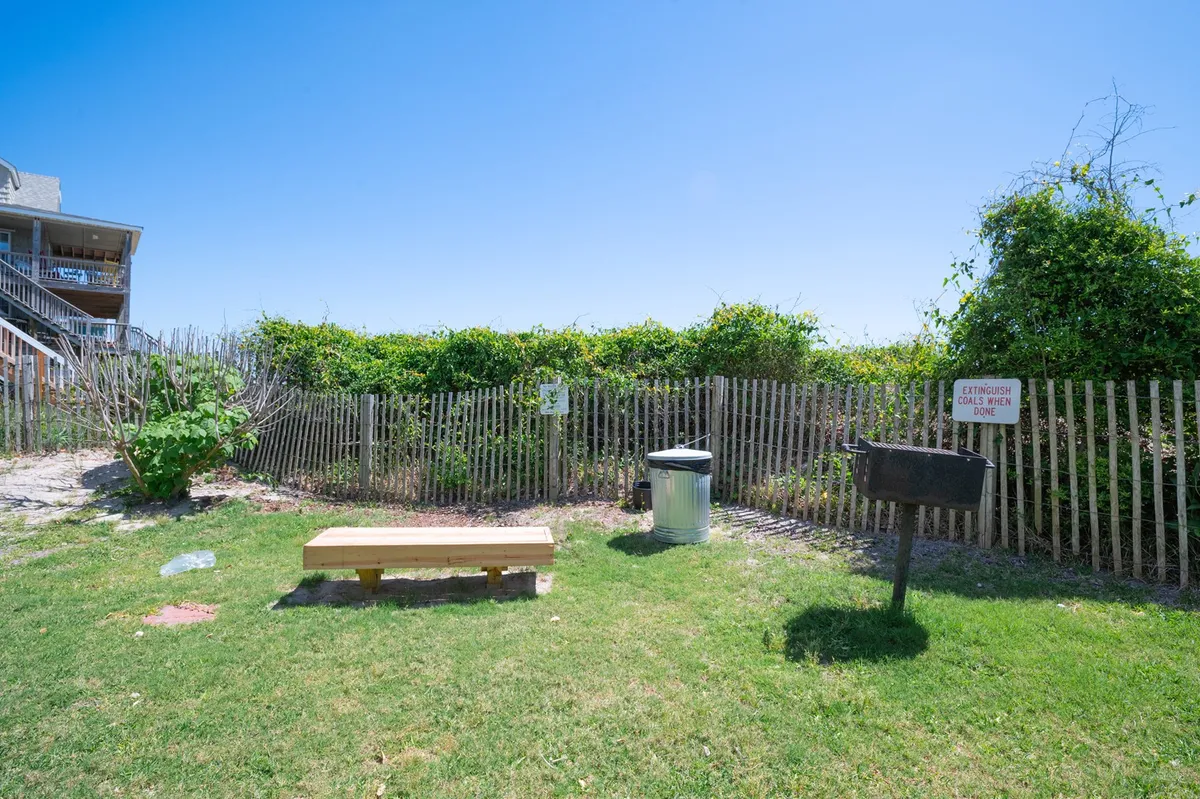 An exterior setting featuring a wooden bench, grill, and lush greenery, creating a relaxed outdoor communal area. The space invites guests to unwind, socialize, and enjoy nature, hinting at the proximity to the beach and opportunities for romantic or social moments outside.