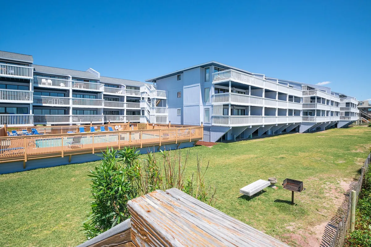 This exterior image captures the central swimming pool, sunlit and inviting, surrounded by grassy areas and overlooked by balconies. Visible in the scene are people relaxing poolside, sunbathing, and a barbecue grill, all contributing to a relaxed, social, resort-style atmosphere.